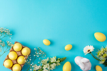 Yellow easter eggs and white spring flowers on blue background with ceramic bunny. Top view, flat...