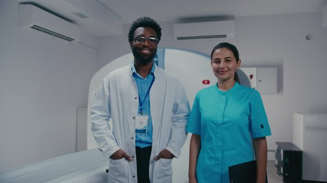 Friendly male doctor with ID badge smiling confidently while standing with hands in pockets. Composed female assistant holding folder and pen beside him after completing patient examination.