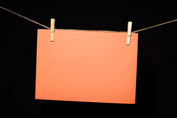 Fotograf&iacute;a de una cartulina naranja sujeta con dos pinzas de madera en una cuerda, sobre un fondo negro. Ideal para dise&ntilde;o gr&aacute;fico, publicidad y presentaciones creativas.