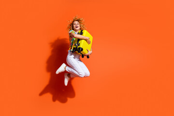Young vibrant woman in trendy casual wear with yellow suitcase jumps against an orange background spreading happiness
