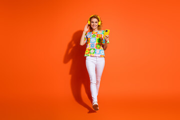Cheerful young woman wearing colorful outfit enjoying music on headphones while holding smartphone against bright orange background.