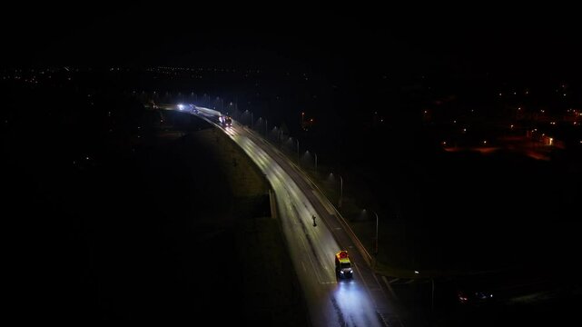 Nighttime flyby shot of a convoy transporting a wind turbine blade along a lit roadway, headlights piercing the darkness as the vehicles move steadily forward.