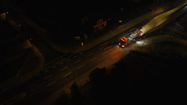 Top-down nighttime shot of a wind turbine blade convoy navigating a residential street, lit by vehicle headlights and streetlights.