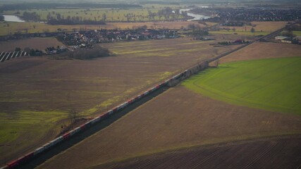 Aerial view of a long freight train passing through fields and countryside, with a rural village and winding river in the background. Movement: follow