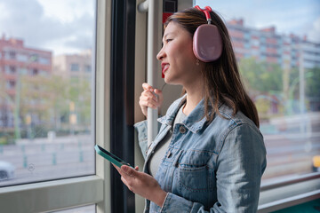 Young Chinese woman in denim jacket holding smartphone and wearing headphones, gazing out tram window. Urban background. Ideal for lifestyle, commuting, travel, music, and technology themes.Copy space