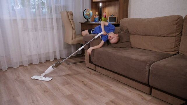 A little boy is fooling around while cleaning the house, he lies upside down on the sofa and cleans the floor with a mop.