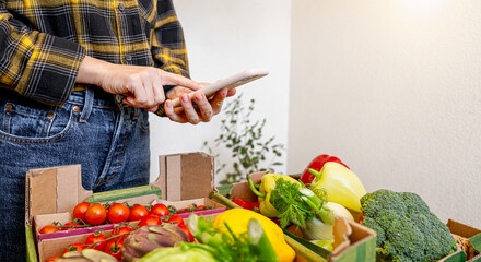 young female farmer preparing online order using smartphone packing organic vegetables in delivery...