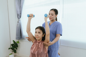 Senior woman exercising with dumbbells during physical therapy in hospital. Physiotherapist helps...