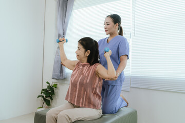 Senior woman exercising with dumbbells during physical therapy in hospital. Physiotherapist helps...