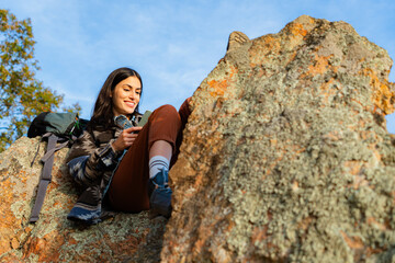 Young woman hiker sitting on a rock, smiling while using her smartphone on a sunny day in the mountains, enjoying nature and adventure