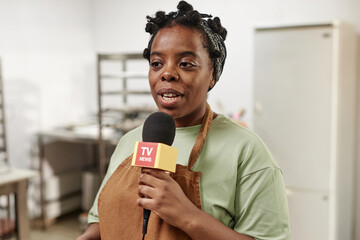 Chest up shot of African American female bakery worker giving interview to journalists speaking into microphone in pastry shop kitchen, copy space