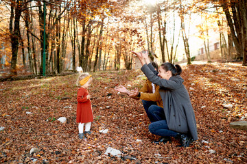Little girl stands in front of mom and dad squatting and throwing up leaves in the autumn park