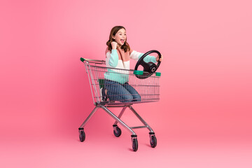 Charming young girl posing in a shopping cart against a pink background, exuding cheerful energy...