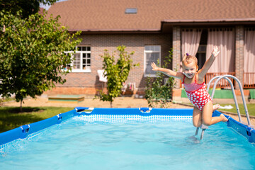 Happy little girl in red swimsuit jumping into outdoor swimming pool at home. Baby girl learning to swim. Water fun for children.