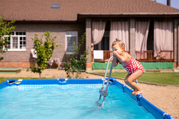 Naklejka premium Little girl in red swimsuit preparing to jump into the pool from the ladder on a sunny summer day