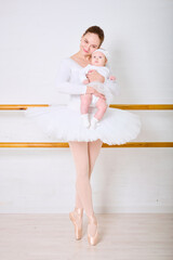 Young woman with brown hair, holding blonde baby girl in white, standing in ballet studio. Both dressed in white, with joyful expressions. Soft lighting, minimal background