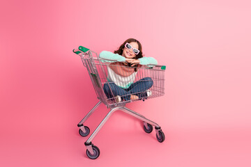 Adorable child in casual outfit enjoying herself while sitting playfully in a shopping cart against...