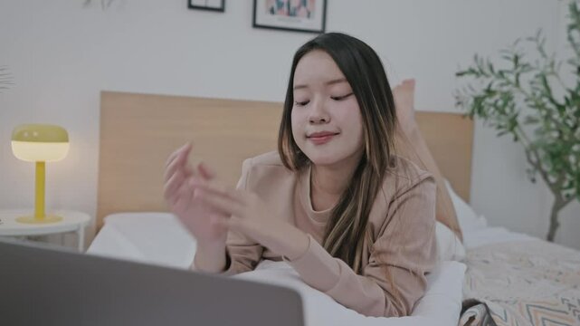 A young woman with long hair enjoys an online conversation on her laptop while nestled in her bed. The bedroom is bright and inviting, with soft lighting and decorative elements.