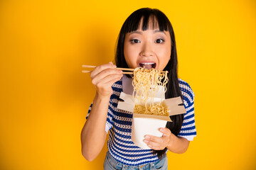 Young cheerful woman enjoying noodles from a takeout box using chopsticks against vibrant yellow...