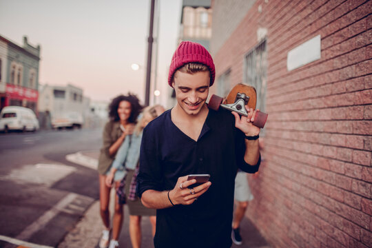 Young skater guy smiling at phone while walking with friends on city street