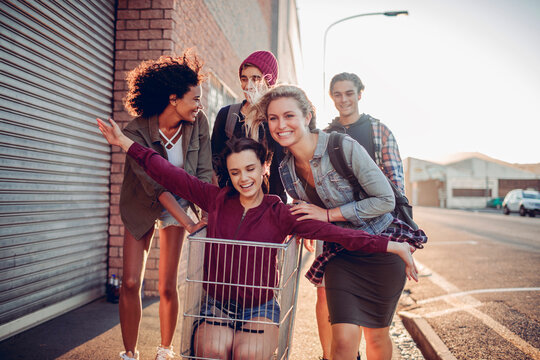 Group of Gen Z friends having fun with drinks and shopping cart on city street at sunset