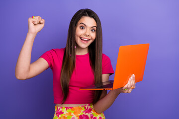 Excited young woman holding an orange laptop and cheering, demonstrating joy and energy against a...