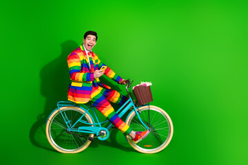 Happy young man in a vibrant rainbow suit riding a colorful bicycle against a green background,...