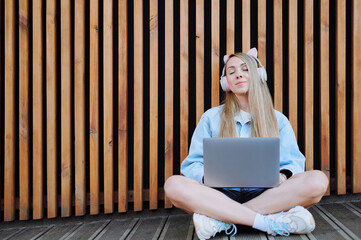 Young woman in denim jacket, cat-ear headphones, eyes closed, working on laptop in cross-legged...