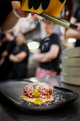 A professional chef grates cheese over a finely plated dish in a restaurant kitchen. The meal consists of seasoned chopped meat with a vibrant green sauce on a creamy base.