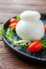 Buratta on a black plate with cherry tomatoes and herbs on a wooden table. Traditional Italian food concept. Vertical orientation. Selective focus.