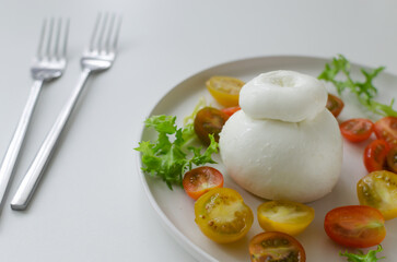 Buratta on a gray plate with cherry tomatoes and herbs on a white table. Traditional Italian food concept. Horizontal orientation. Selective focus. Copy space