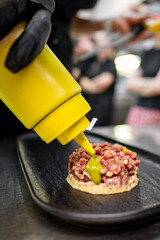 A gloved hand squeezes a yellow bottle, releasing green sauce onto a finely chopped meat dish served on a black plate. The background is blurred, showing chefs working in a professional kitchen