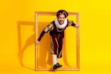 Playful young man in noble costume posing with picture frame against vibrant yellow background