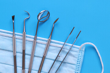Dental mirror, tweezers and several hooks on disposable medical mask. Collection of dental instruments on blue background. Diagnostic dental instrument set. Oral and dental health concept. Medicine