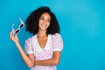 Smiling young woman holding stylish pink sunglasses against a vibrant blue background, radiating...