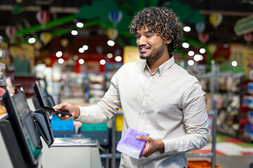 A smiling man uses contactless payment at a grocery store checkout. He holds his phone near the payment terminal and a package of groceries.