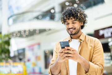 A smiling man with curly hair looks at his phone in a shopping mall, enjoying his smartphone in public.
