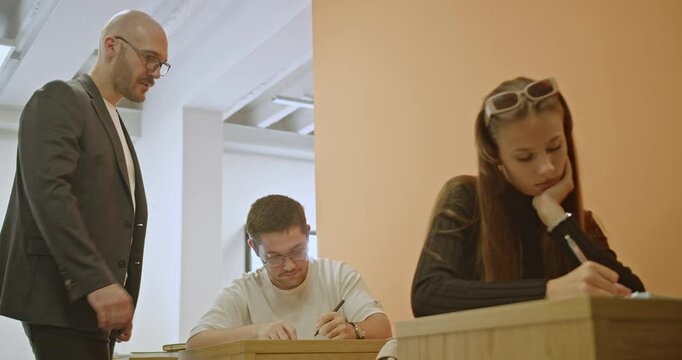 High school students focused during an exam, with their professor providing assistance as needed.