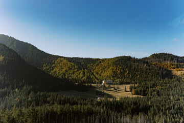 A scenic view of a mountain valley covered in evergreen and golden autumn trees. A solitary house sits in the open meadow, surrounded by forested hills under a clear blue sky.