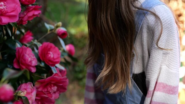 Young woman with long hair in denim dungarees. European woman smelling camellia flowers in the garden. Hand holding red flowers. Multiple outdoor shots.