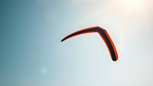 Red wooden boomerang flying through blue sky in sunlight, captured mid-air for dynamic motion effect.  