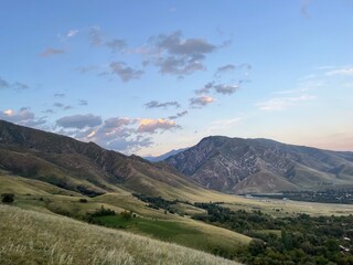 serene mountain valley at sunset with rolling green hills, dramatic rocky peaks, and a small village nestled in the landscape

