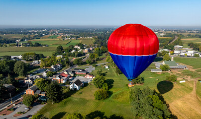 A vibrant hot air balloon glides gracefully above lush green fields and quaint homes in a...