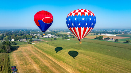 Obraz premium Two vibrant hot air balloons float gracefully above an expansive green field dotted with farms. The clear blue sky enhances the serene morning atmosphere.