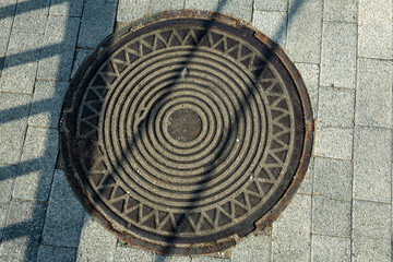 Old manhole cover on light paved street. City infrastructure. Warm summer day.