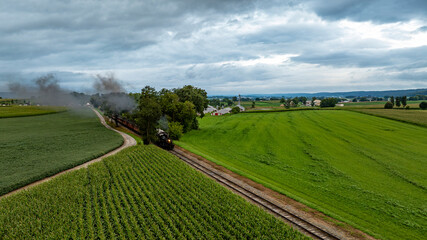 Obraz premium A vintage steam train moves along a meandering railway, surrounded by vast green fields and under a cloudy sky. The landscape captures the essence of rural tranquility.