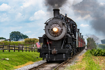 A historic steam locomotive travels along rural tracks, surrounded by lush greenery and waving American flags. The vibrant sky adds to the charm of this nostalgic journey.