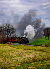 A vintage steam locomotive billows dark smoke as it travels across lush green fields. The overcast sky adds a dramatic backdrop to this nostalgic journey.