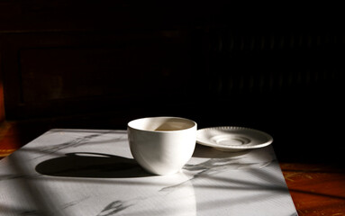 Coffee cup and blur coaster place on table with strong shadow and black space. Still-life white cup in cozy sunny day. Art style, abstract, warm, decor, fresh drink, minimal, relax, darkness concepts.