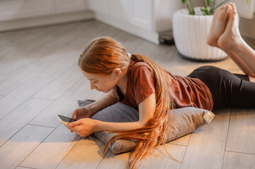 A red-haired girl with long hair is lying on the kitchen floor playing and scrolling her phone....